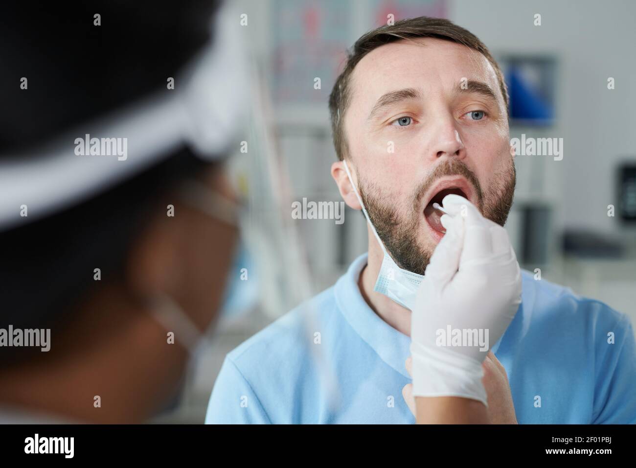 Face of young male patient being tested for covid with oral swab held by gloved female nurse or clinician while sitting in front of her Stock Photo
