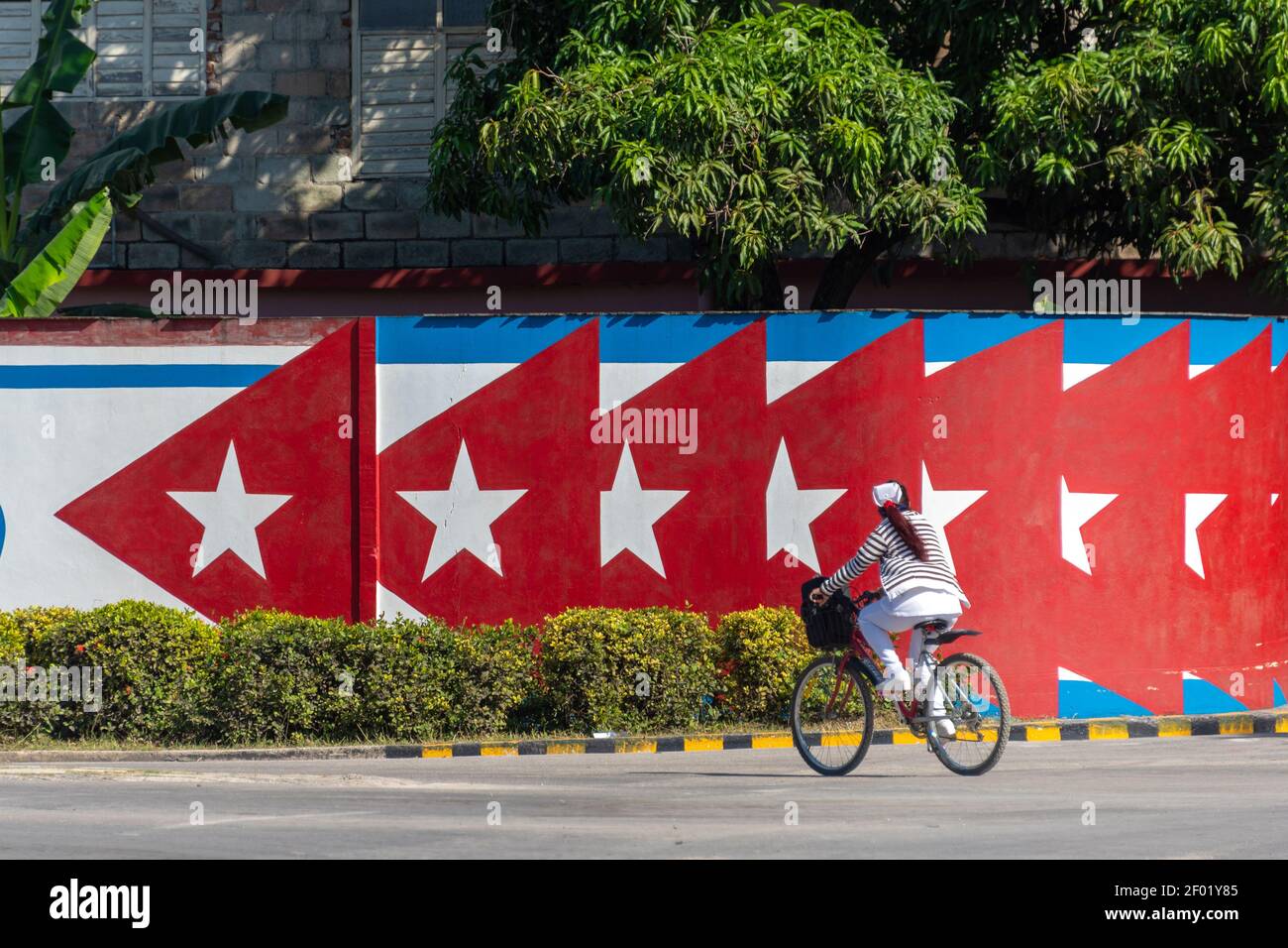 Woman nurse rides a bicycle by a fence with a Cuban flag decoration, Holguin, Cuba Stock Photo