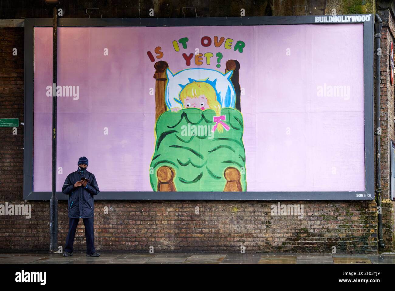 London, UK. - 17 Feb 2021: A face-masked pedestrian stands in front of a humourous fly-posted billboard questioning whether the COVID-19 coronavirus pandemic is over yet. Stock Photo
