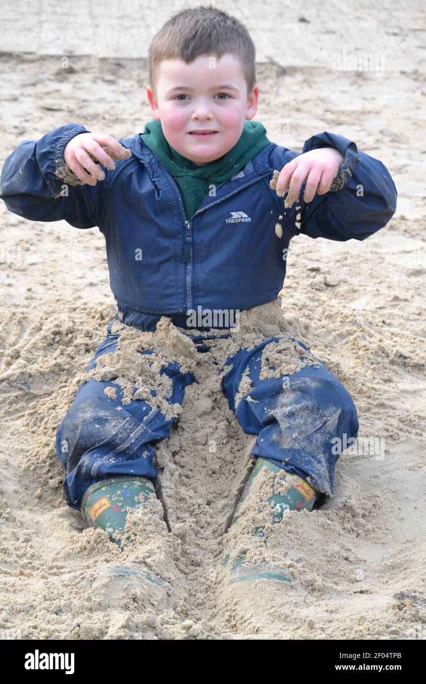 Young Child Playing In The Sand On A Winters Day - Primary School Aged - All In One Waterproofs - Smiling - Young Lad - At The Park - Playground - UK Stock Photo