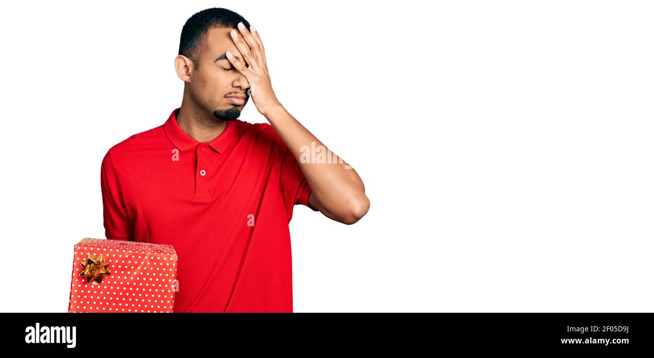 Young african american man holding gift stressed and frustrated with hand on head, surprised and angry face Stock Photo