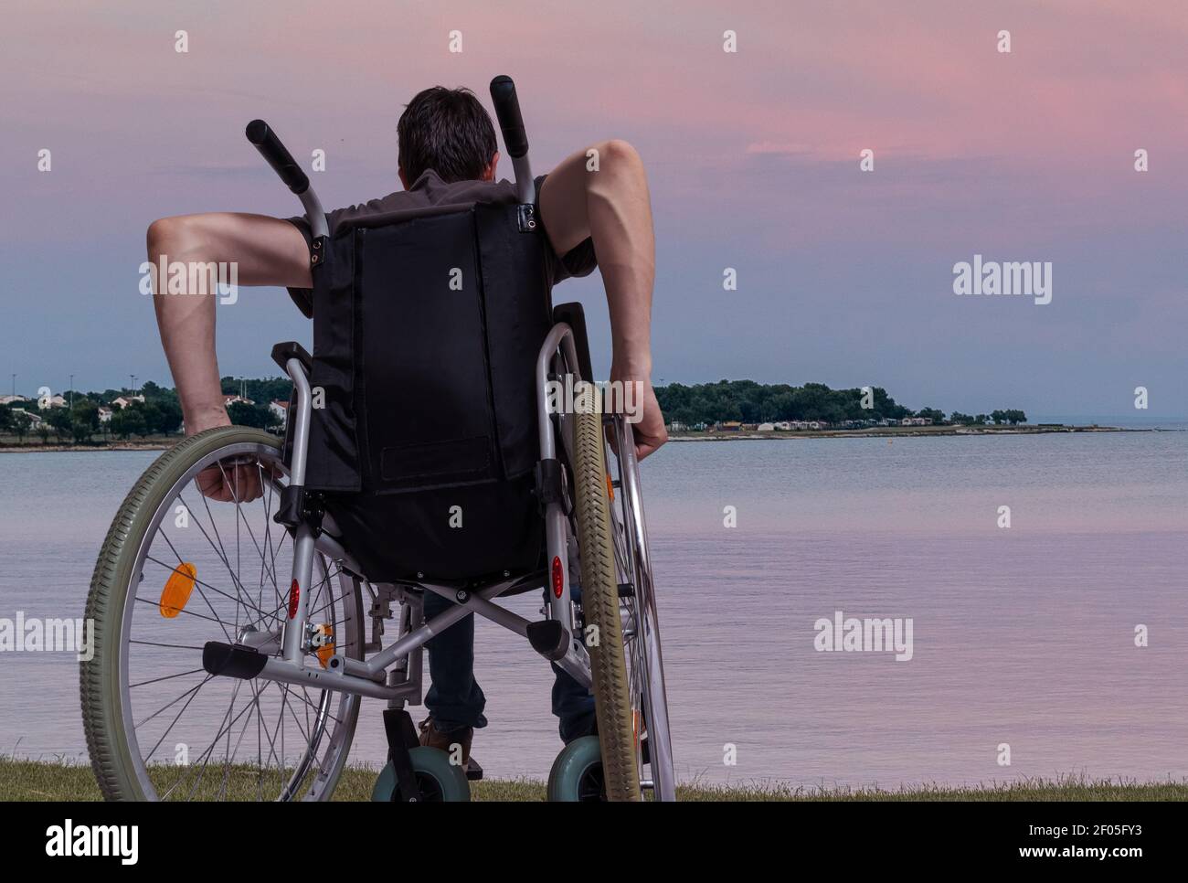 Young man is sitting on wheelchair near sea at sunset. Stock Photo