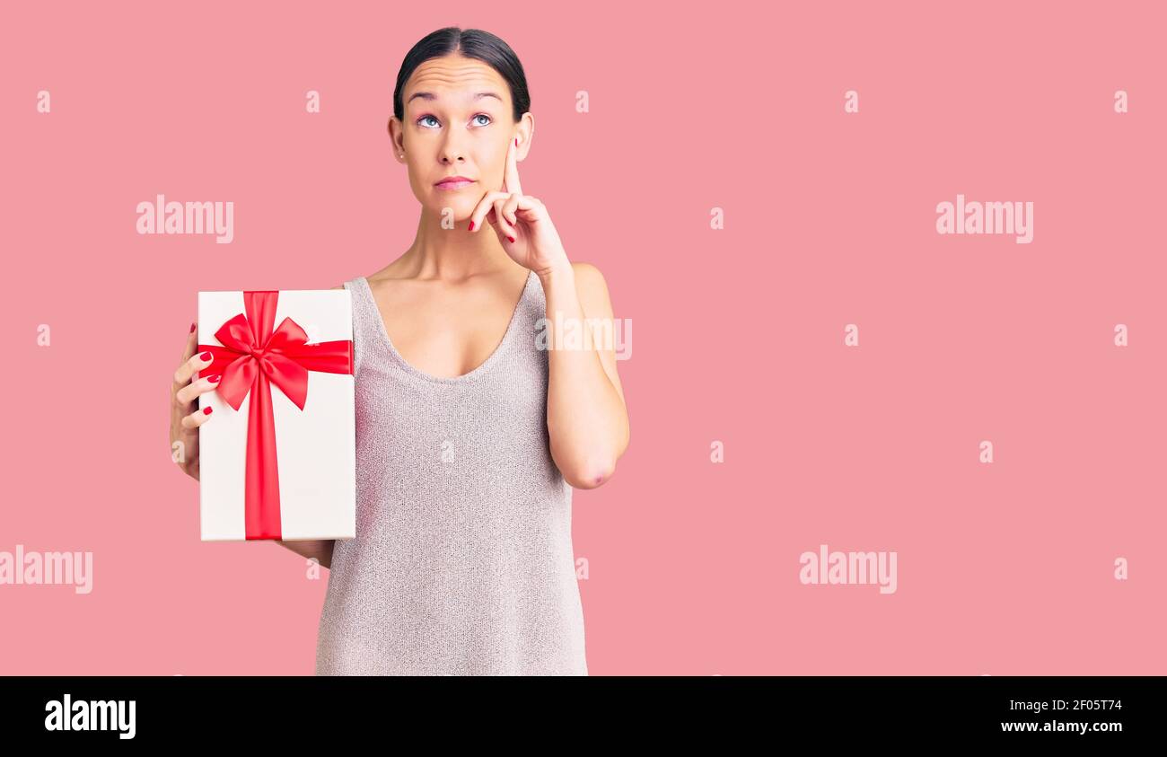 Beautiful brunette young woman holding gift serious face thinking about question with hand on chin, thoughtful about confusing idea Stock Photo
