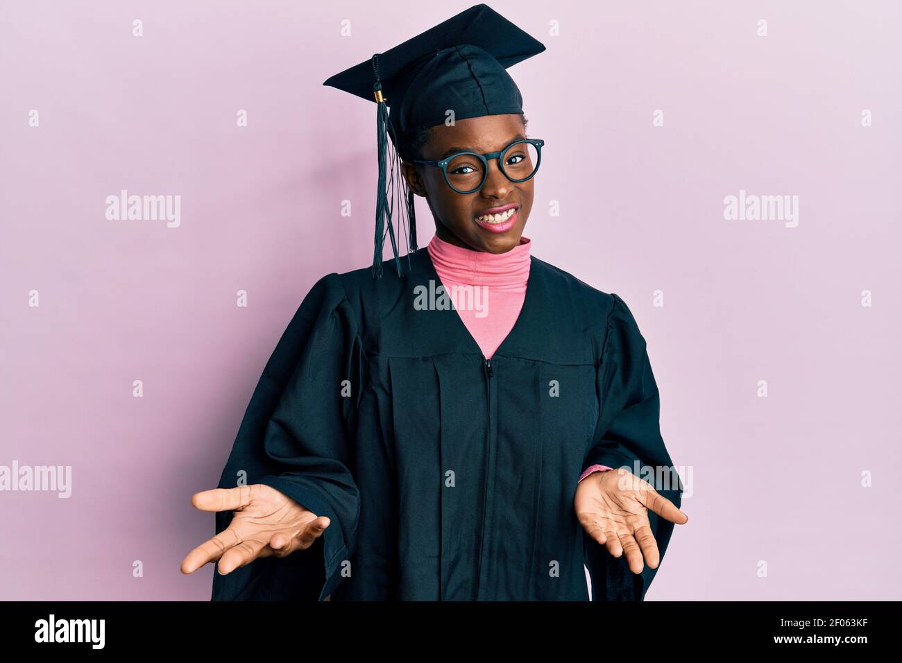 Young african american girl wearing graduation cap and ceremony robe smiling cheerful with open arms as friendly welcome, positive and confident greet Stock Photo