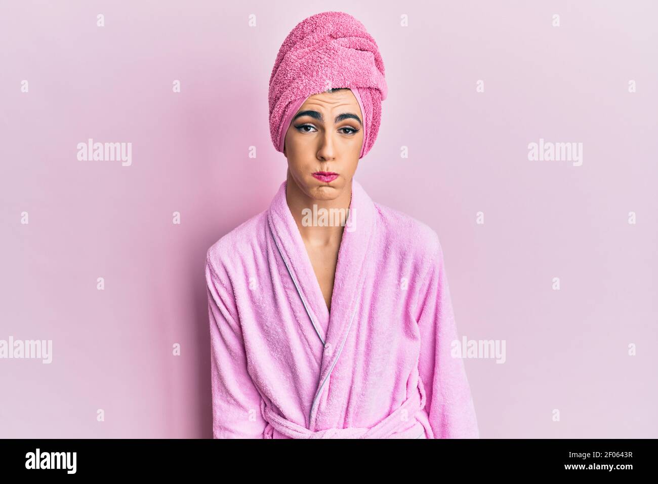 Young man wearing woman make up wearing shower towel on head and bathrobe puffing cheeks with funny face. mouth inflated with air, crazy expression. Stock Photo