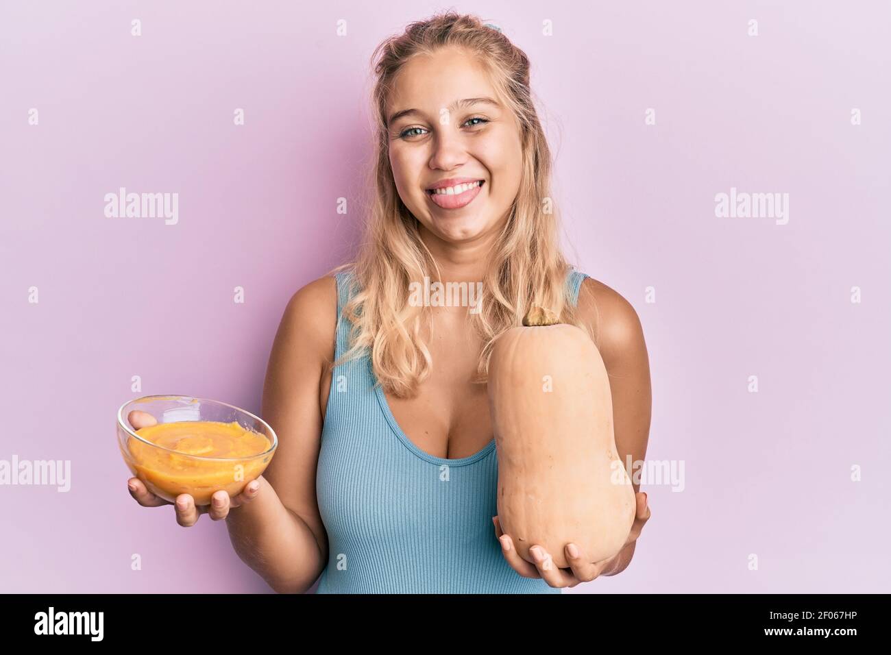 Young blonde girl holding fresh pumpkin and soup sticking tongue out happy with funny expression. Stock Photo
