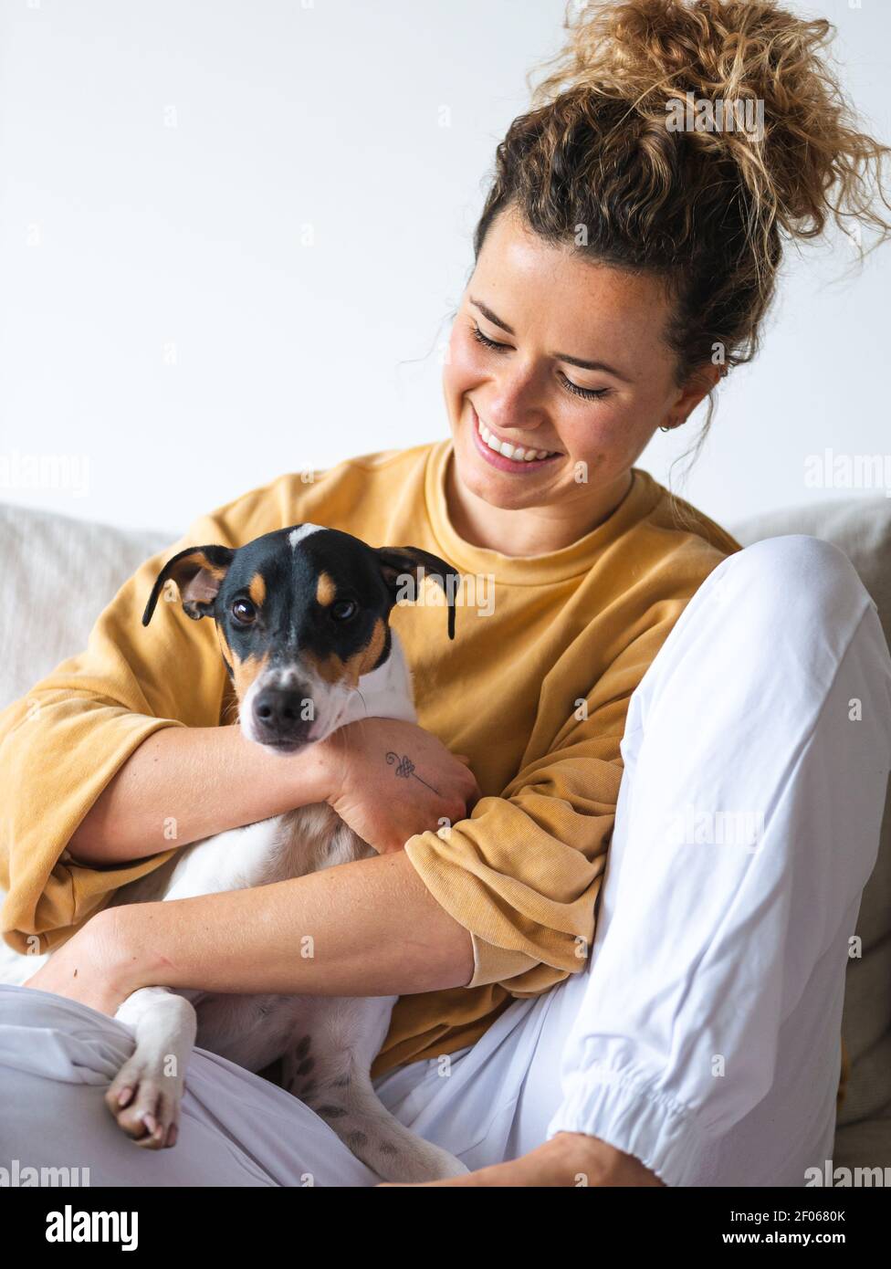 Cheerful Woman with curly hair an casual clothes sitting with obedient Ratonero Bodeguero Andaluz dog on sofa in light apartment Stock Photo