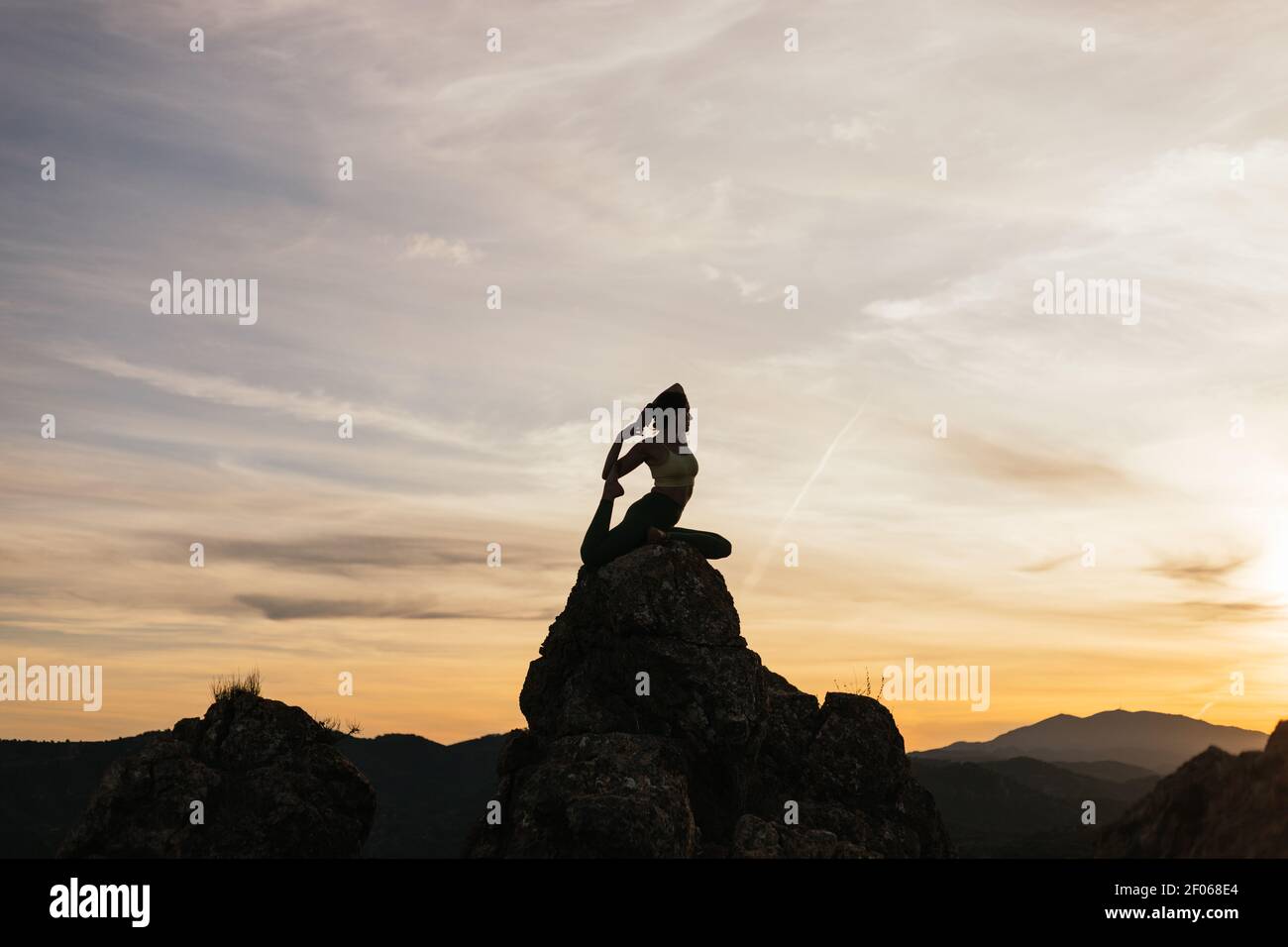 Silhouette of slim female performing One Legged King Pigeon pose on top of mountain at bright sundown Stock Photo