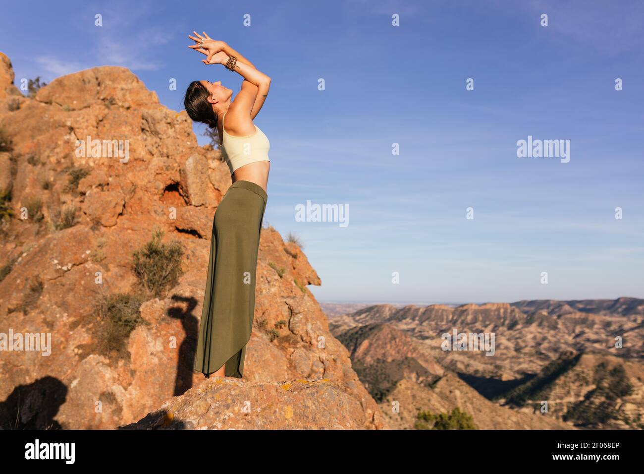 Side view of slim female in casual outfit performing Mountain with Arms Up and Backbend posture on slope of rocky mountain Stock Photo