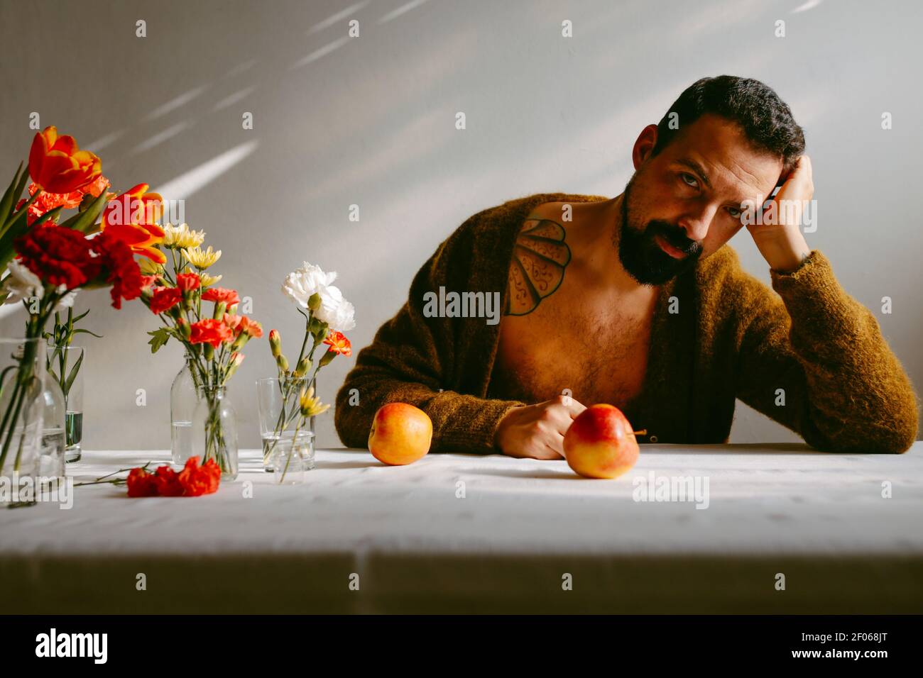 Bearded pensive male leaning on hand sitting at table with ripe apples and flowers in glasses Stock Photo