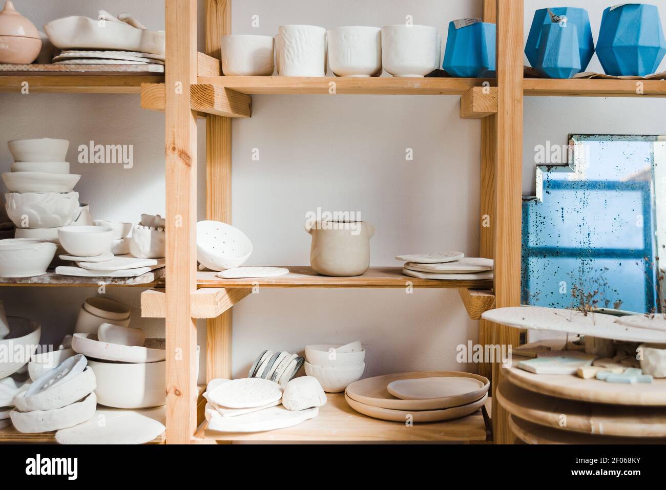 Collection of handmade ceramic bowls and vases with pots and plates with old mirror near different types of utensil standing on wooden shelves in ligh Stock Photo