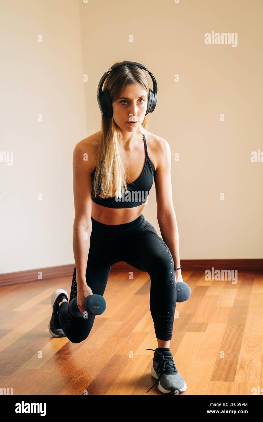Full body of determined female in sportswear with headphones listening to music while doing lunges exercise with weight at home Stock Photo
