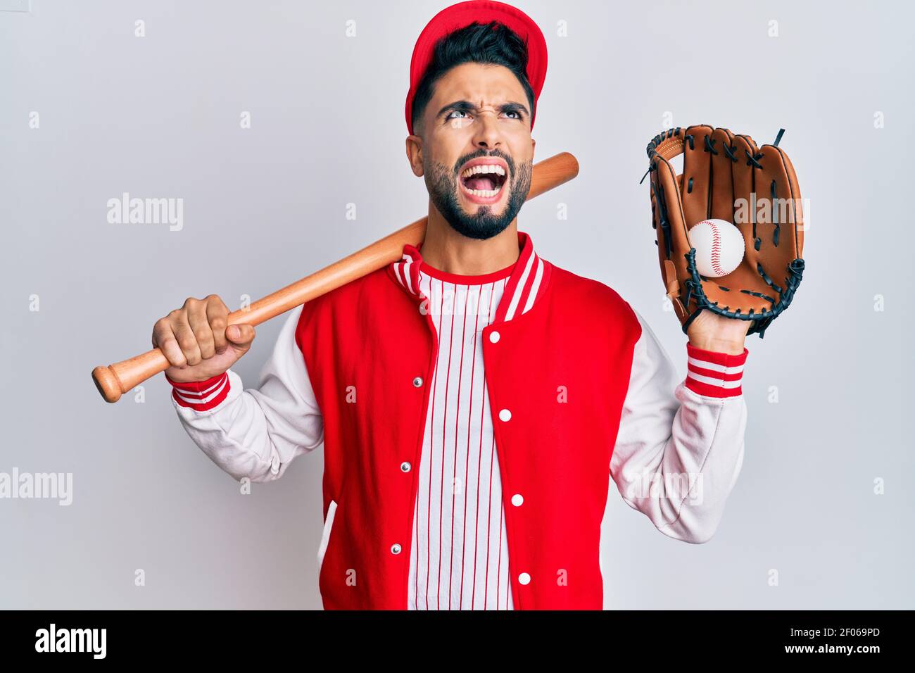 Young man with beard playing baseball holding bat and ball angry and