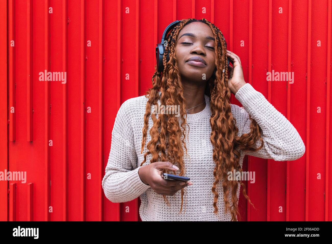 Positive serene African American female in wireless headphones enjoying songs  with closed eyes while listening to music on street against red wall Stock  Photo - Alamy