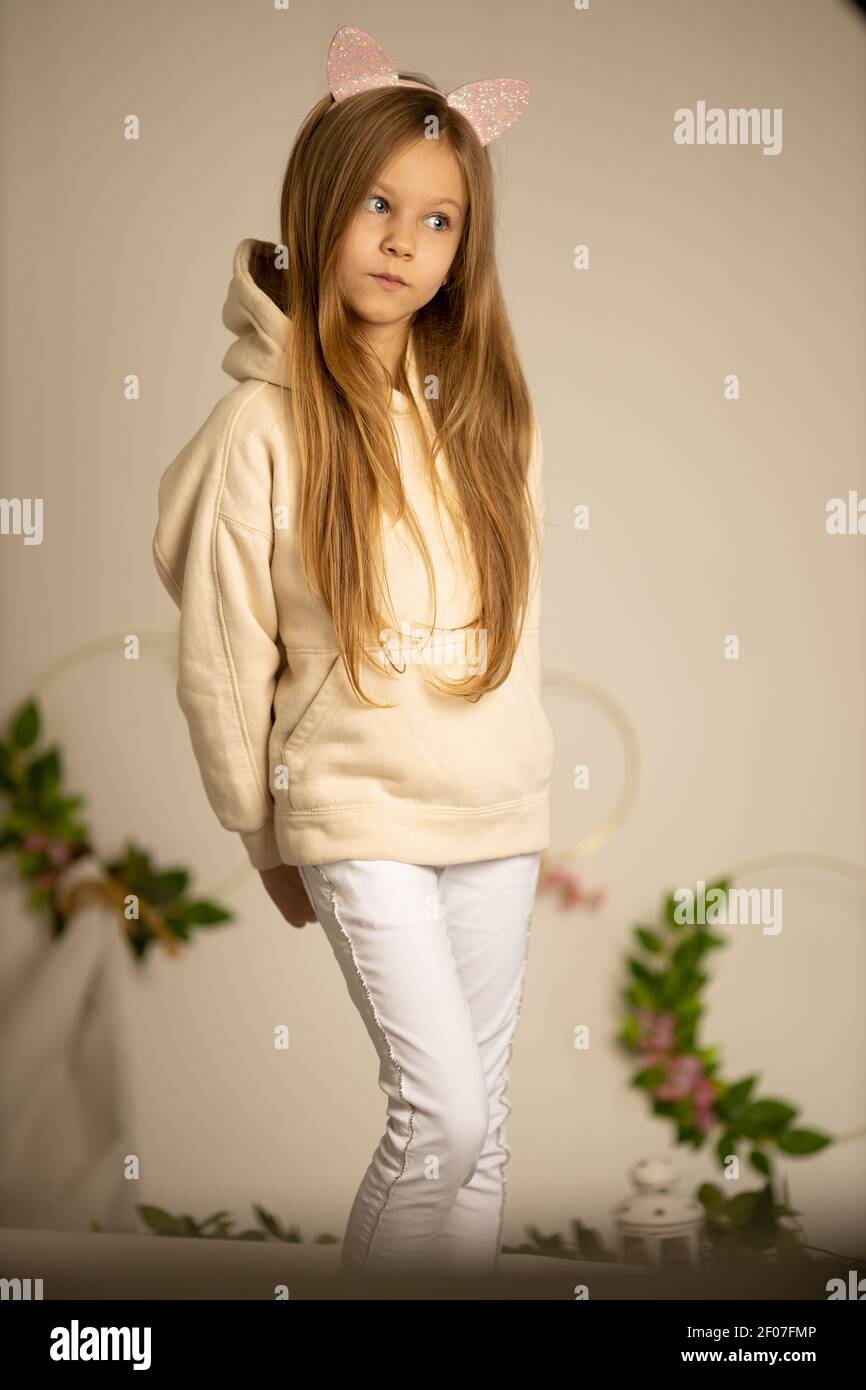 pensive girl 8-9 years old in a beige sweatshirt and white pants, holding her hands behind a bed on a cream background Stock Photo