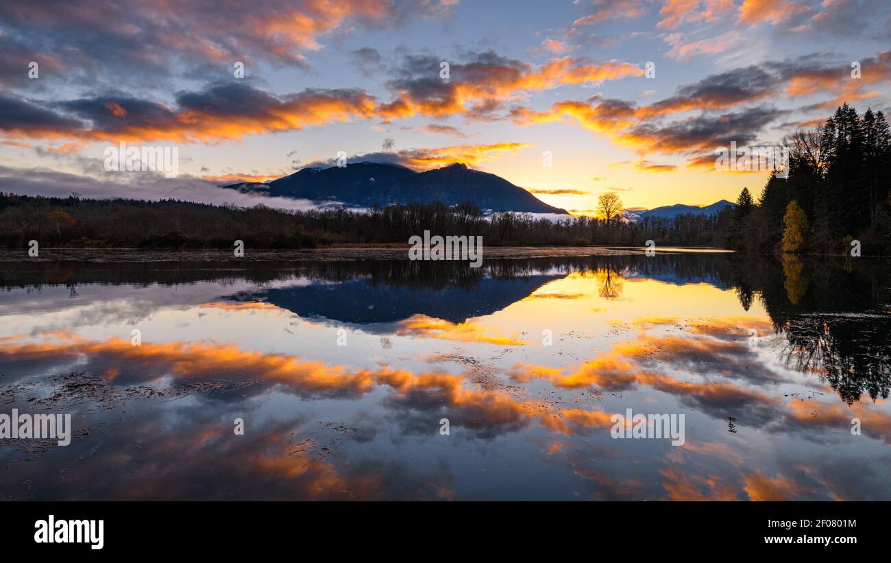 Mount Si reflects at dawn in Borst Lake as the rising sun adds colors to a sparse collection of morning clouds above the Cascade Mountains Stock Photo