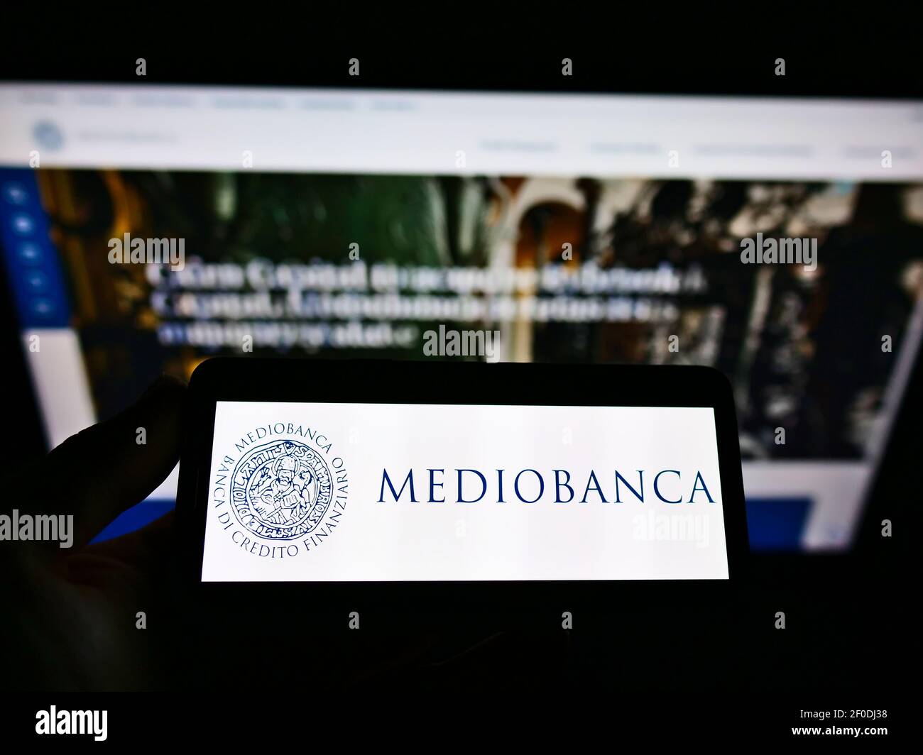 Person holding mobile phone with company logo of Italian investment bank Mediobanca SpA. on screen in front of web page. Focus on cellphone display. Stock Photo