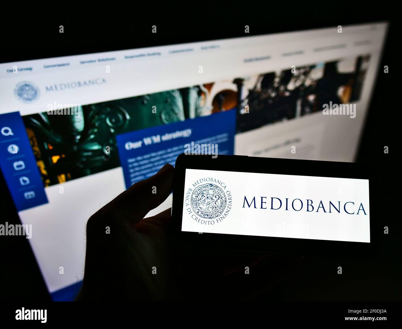 Person holding smartphone with logo of Italian investment bank Mediobanca S.p.A. on screen in front of website. Focus on phone display. Stock Photo