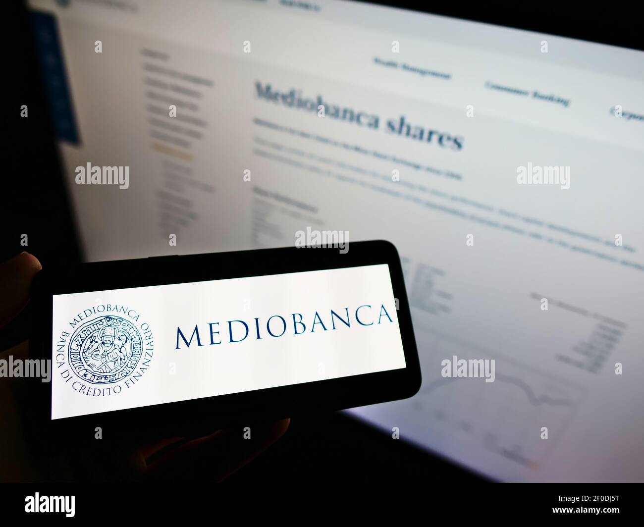 Person holding cellphone with business logo of Italian bank Mediobanca S.p.A. on screen in front of company webpage. Focus on phone display. Stock Photo