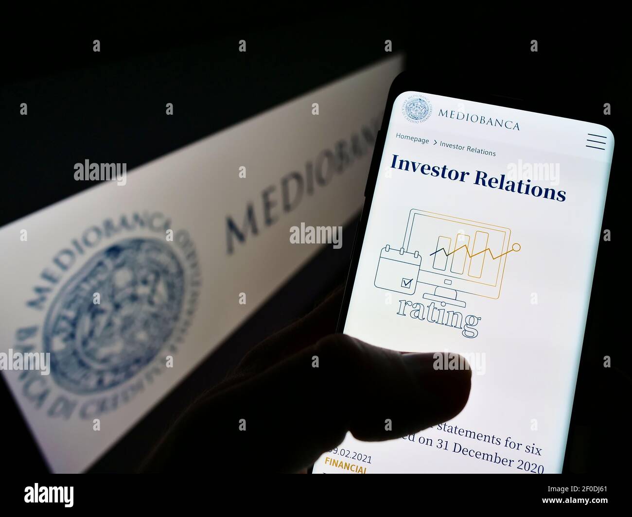 Person holding mobile phone with website of Italian investment bank Mediobanca SpA. on screen in front of logo. Focus on center of phone display. Stock Photo