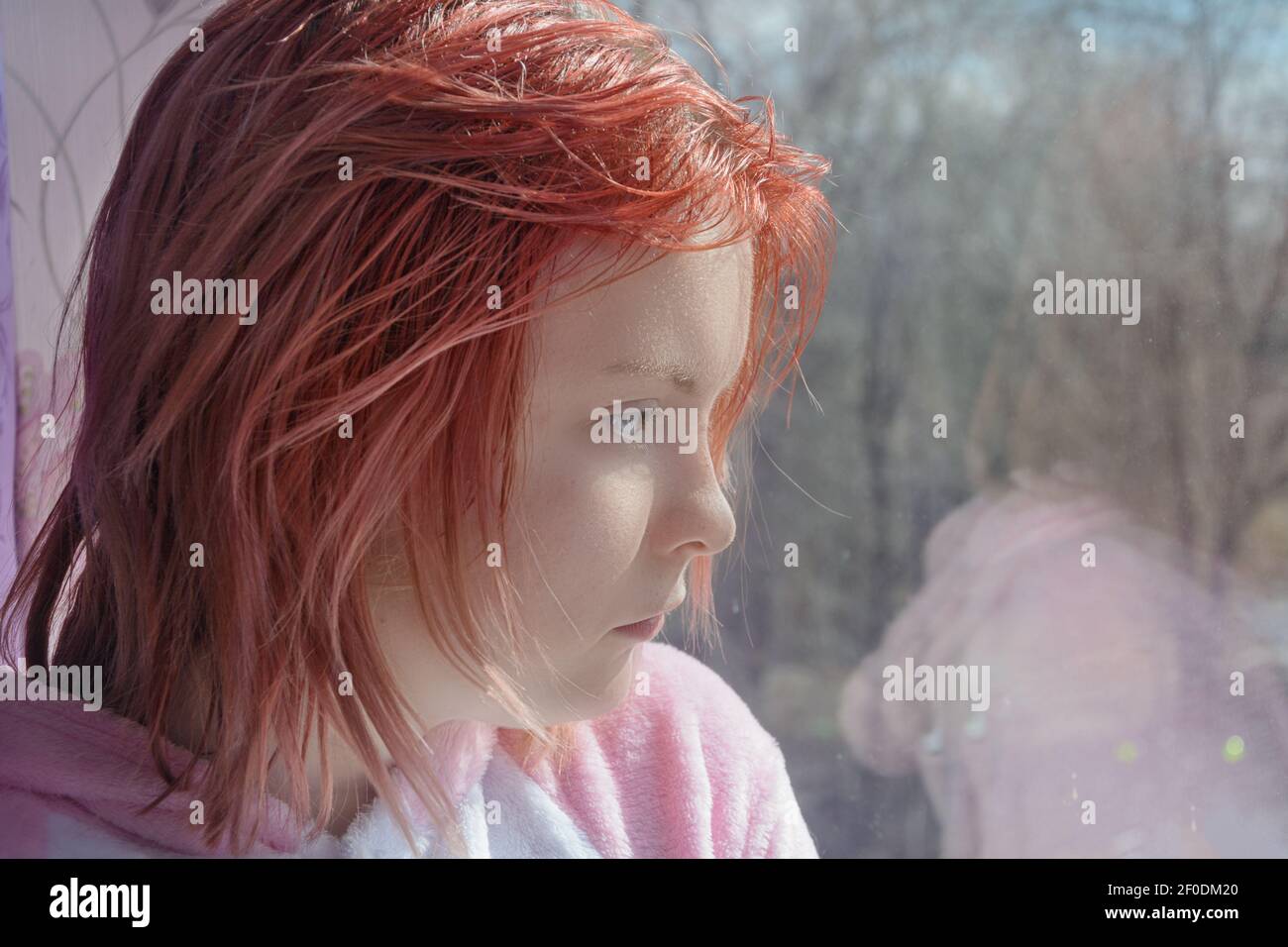 Teenage girl with red hair looks out window to street. Sad emotions on face. Close-up, selective focus. Stock Photo