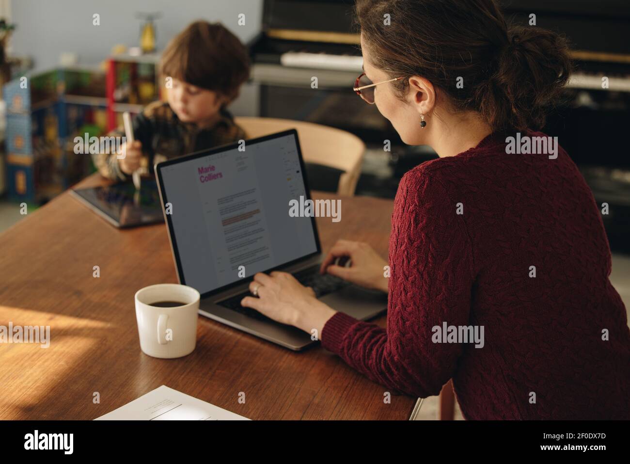 Woman working on laptop with her son using digital tablet in background. Working mother with son playing with tablet computer. Stock Photo