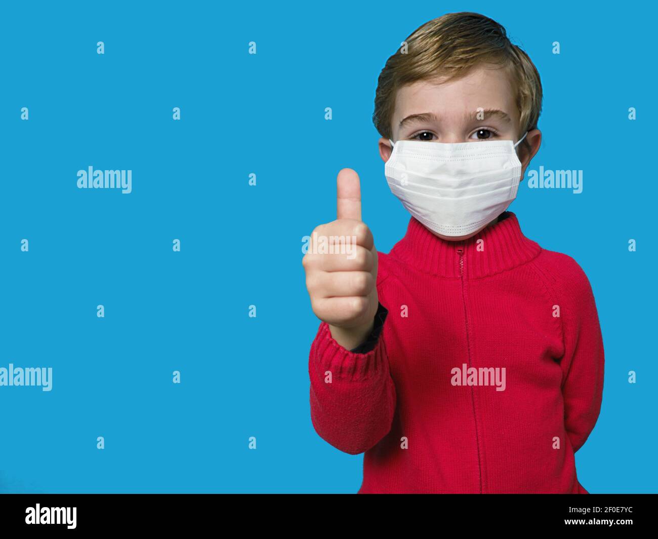 Child with white face mask show thumb up on blue background. Covid-19 coronavirus pandemic Stock Photo