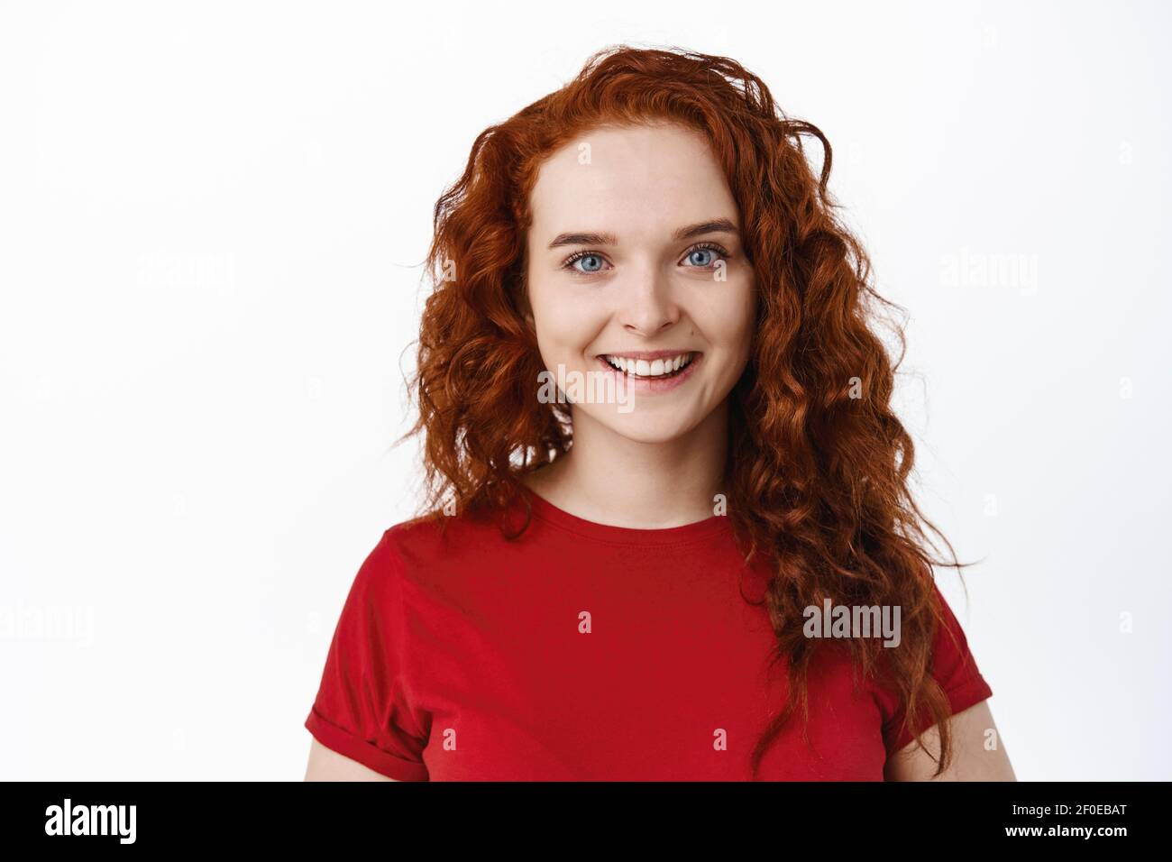Close-up of candid smiling woman with red curly hair and pale healthy skin, looking cheerful at camera, standing in t-shirt against white background Stock Photo
