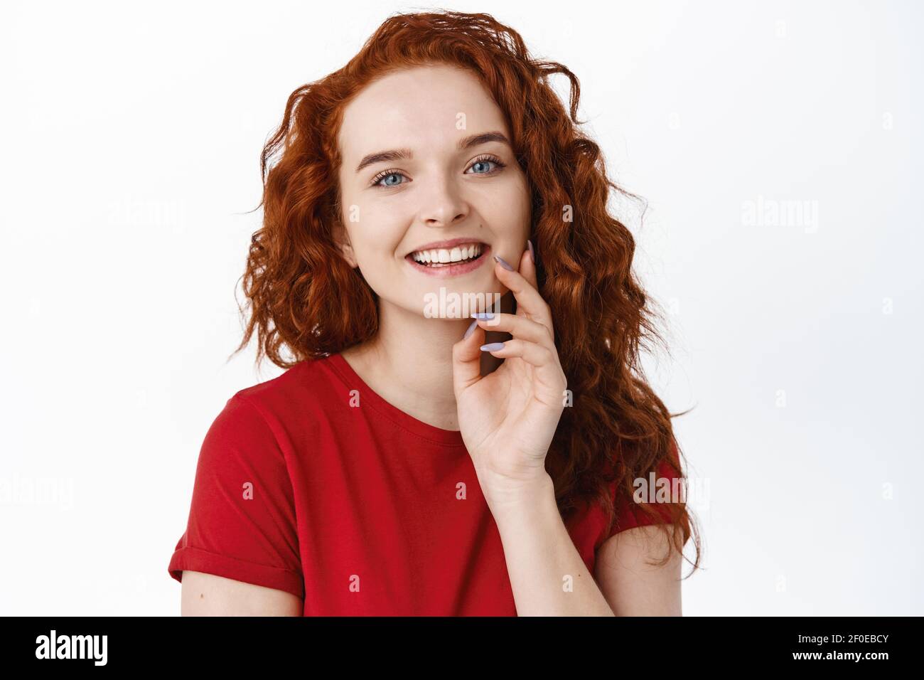 Close up portrait of smiling happy woman with ginger curly hair, touching pale smooth and healthy skin with fingertips, looking cheerful and laughing Stock Photo