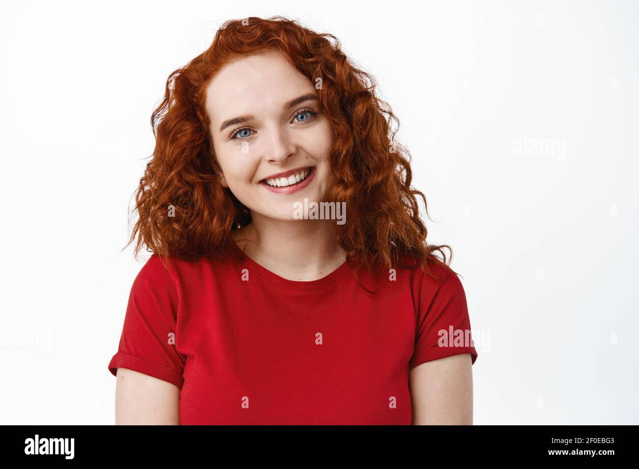 Portrait of cute teen girl with red curly hair and pale natural skin without make up, tilt head and smile at camera friendly, standing over white Stock Photo