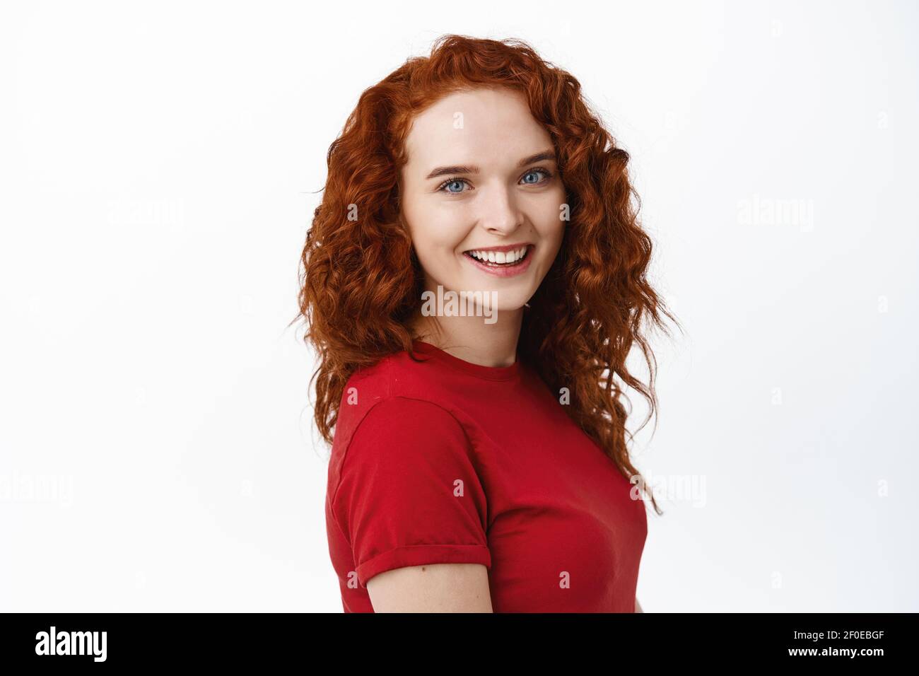 Portrait of confident young pretty woman with curly red hair, natural pale skin and light make-up, showing her candid white smile and looking at Stock Photo