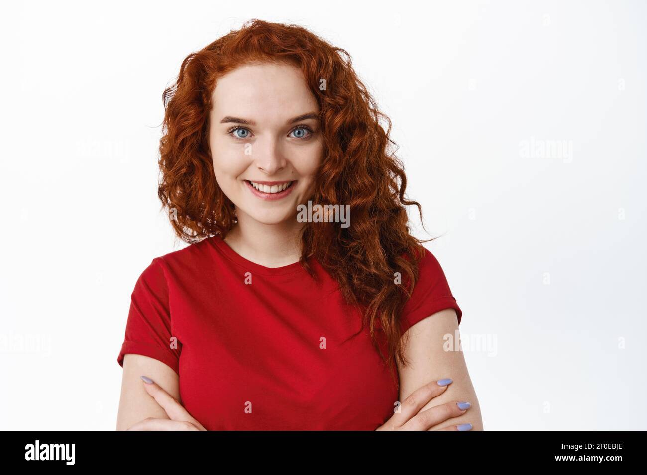 Close-up portrait of confident woman with ginger curly hair, cross arms on chest and smiling, have pale skin without blemishes and blue eyes, white Stock Photo