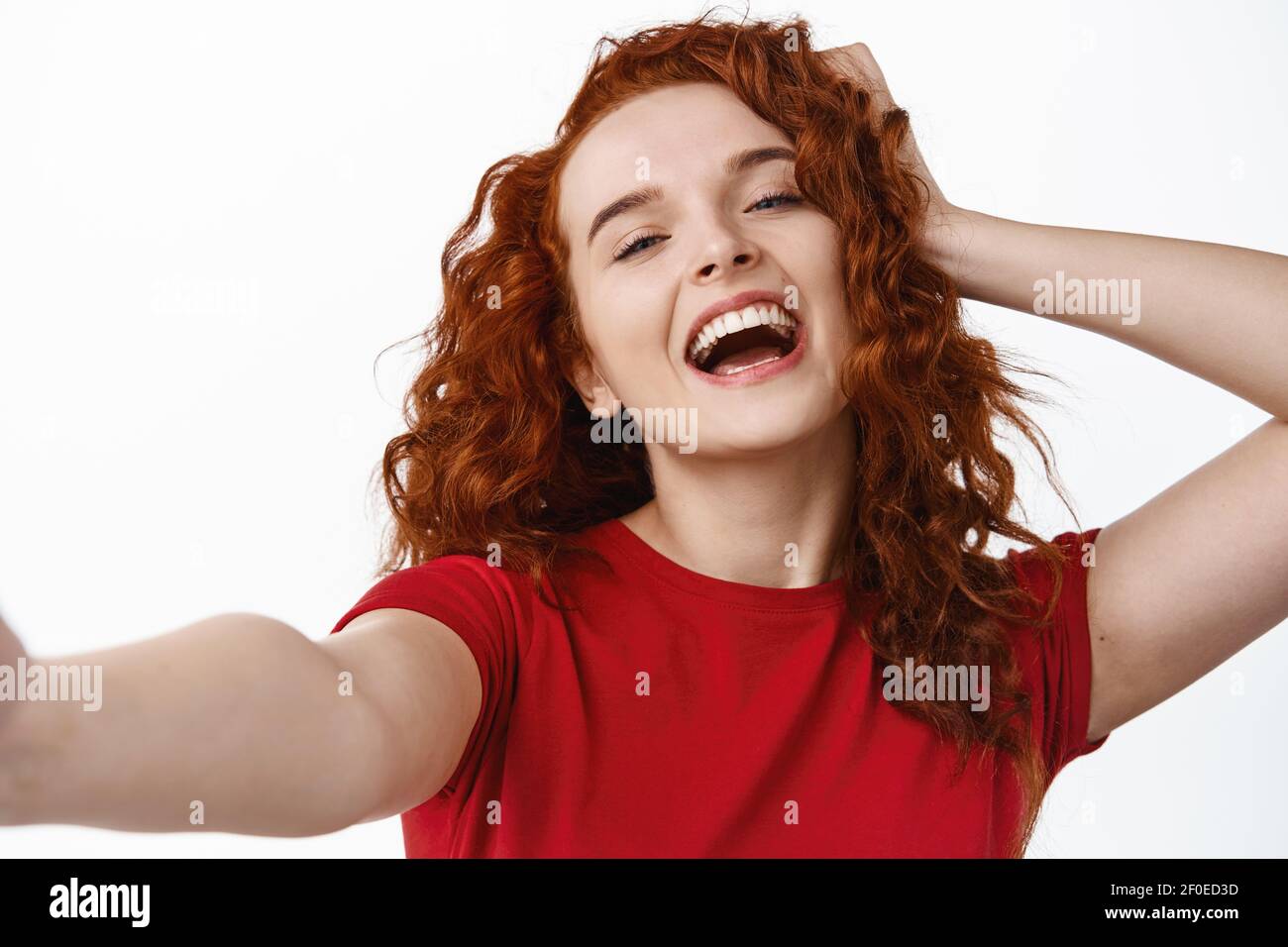 Close up portrait of carefree and happy ginger girl, touching her curly natural hair and laughing while taking selfie, holding camera with hand, take Stock Photo