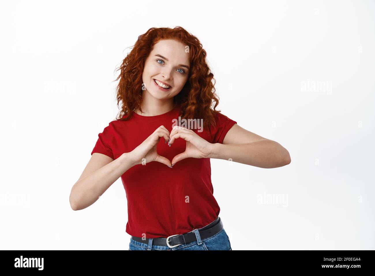 Portrait of beautiful ginger girl with curly hair and pale skin, showing heart I love you gesture near chest, express like or sympathy, standing Stock Photo