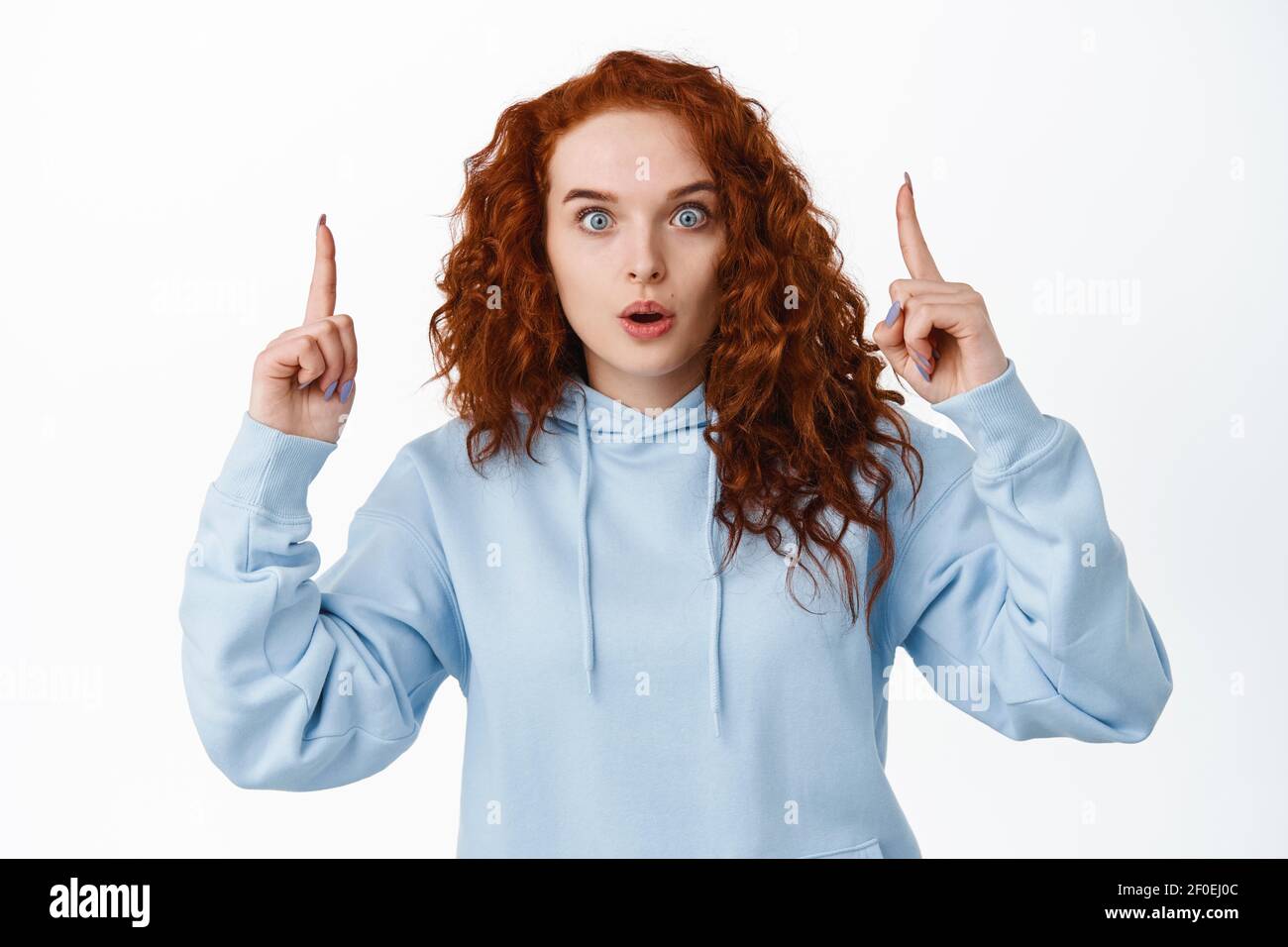Impressed redhead girl stare at camera surprised and gasping, pointing fingers up at super cool thing, standing against white background in casual Stock Photo