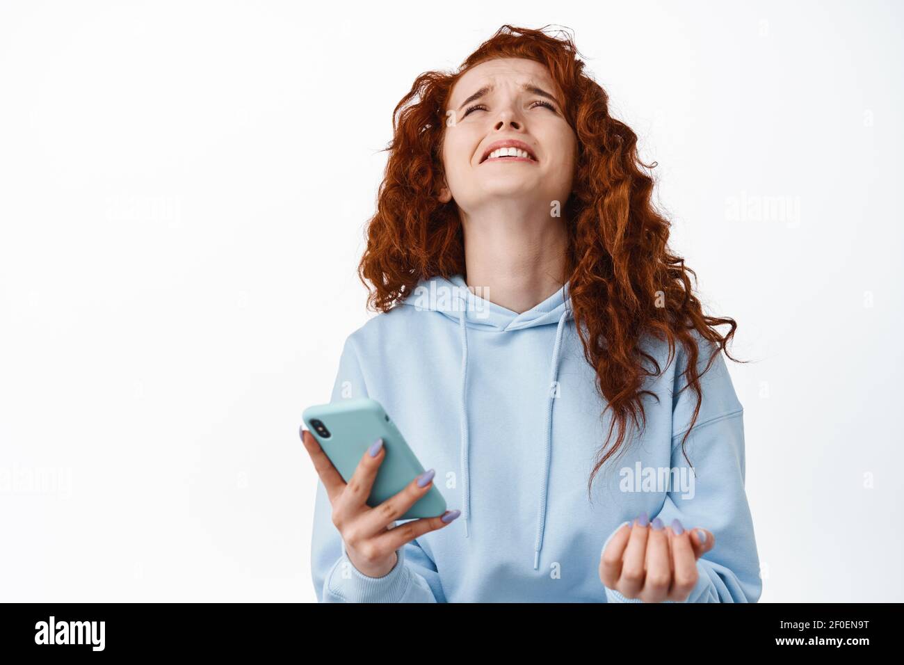 God why me. Miserable and sad redhead girl complain on unfair and cruel life, holding smartphone, raising head up and whining as losing online Stock Photo