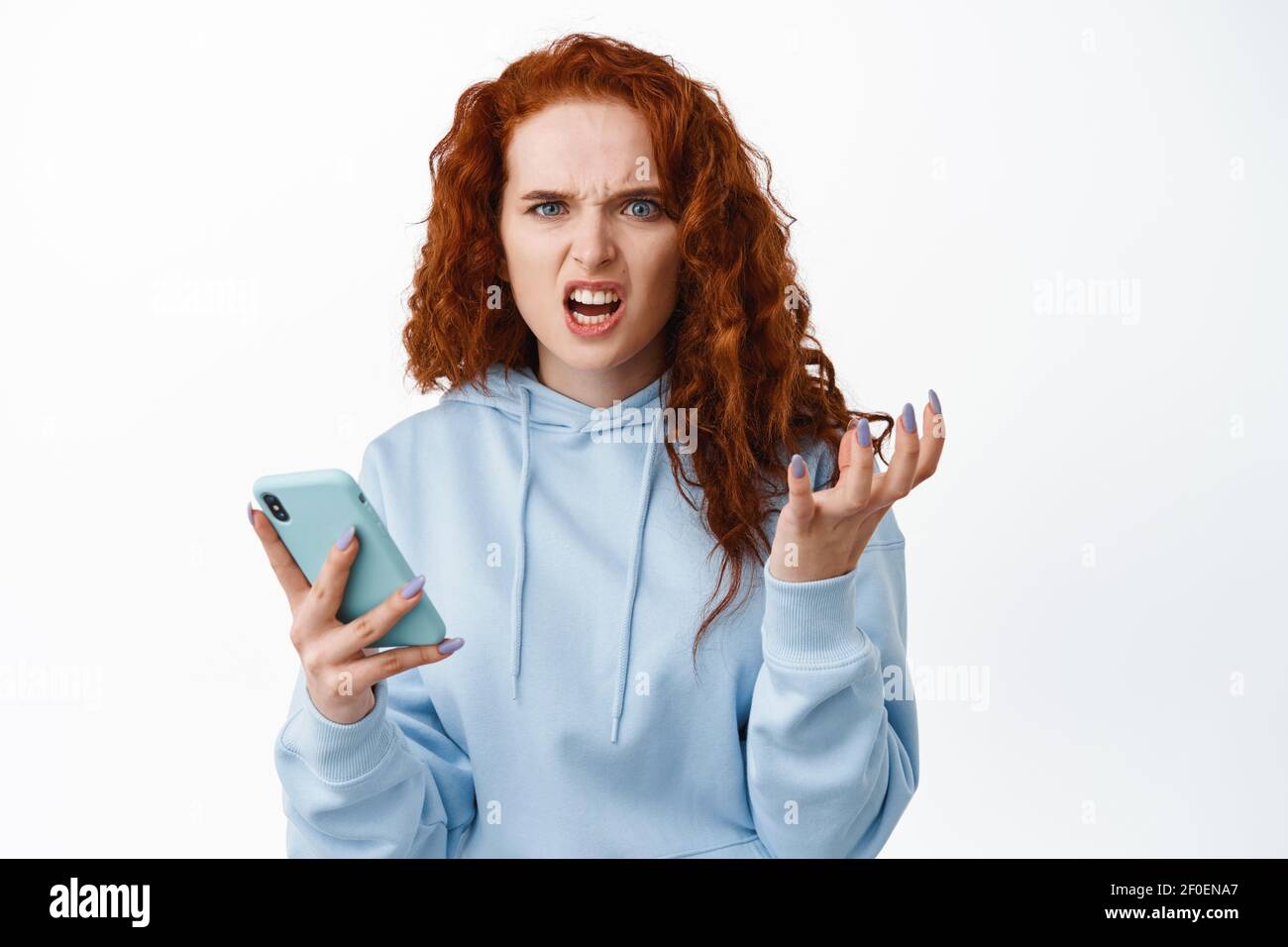 Angry and annoyed redhead girl holding smartphone, frowning and grimacing mad, complaining at something bothering, being dumped by message, white Stock Photo