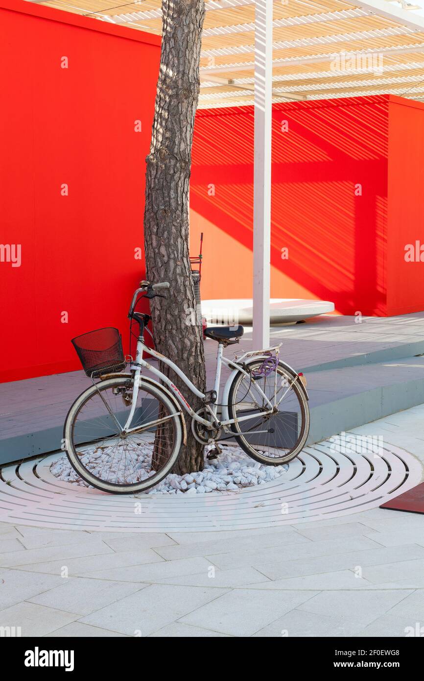 Bicycle leaning against a tree  with white pebbles and circular boards in front of a bright red wall  on Lido Island, Venice, Veneto, Italy. Tradition Stock Photo
