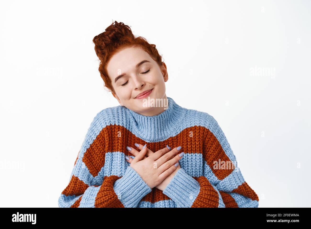 Close up of beautiful ginger girl with combed hair and pale skin, smiling with eyes closed and hands on heart, dreaming about lovely memory, standing Stock Photo