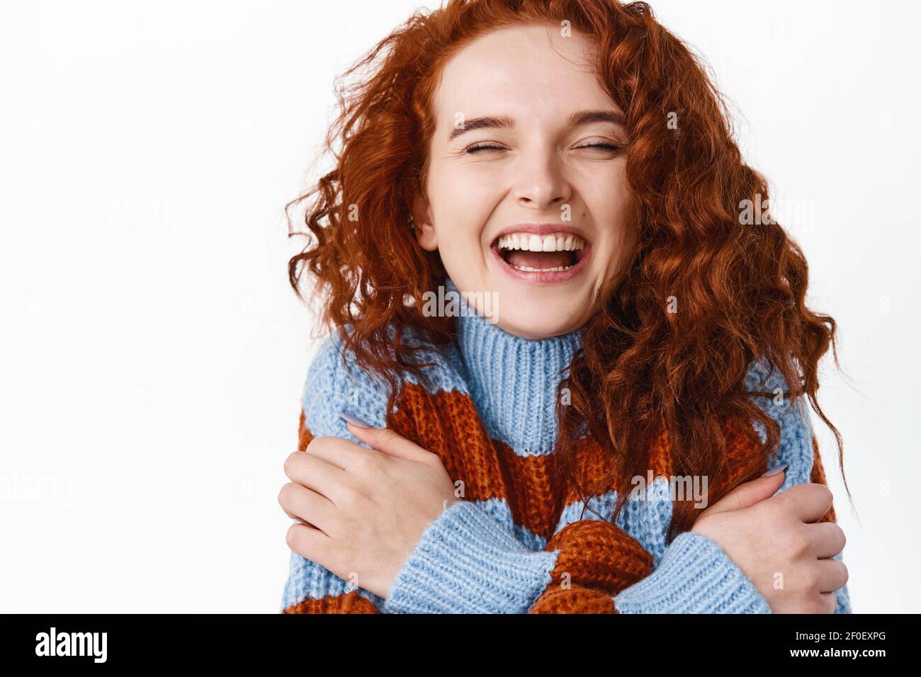 Close up portrait of happy beautiful redhead woman with curly natural hair, pale smooth skin, laughing sincere with eyes closed and hugging herself Stock Photo