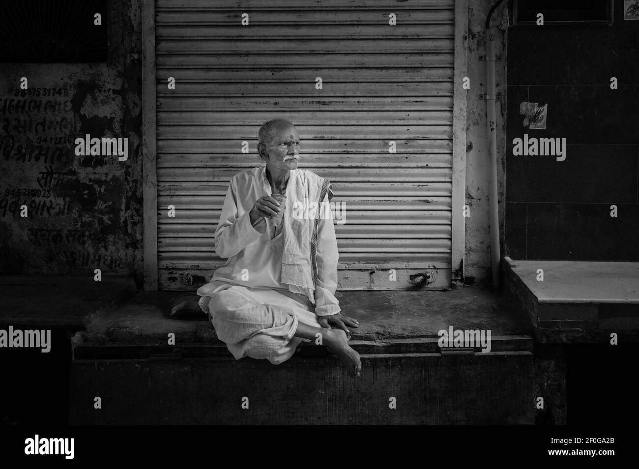Candid portrait of older man sitting cross-legged looking thoughtful and drinking tea in street scene in Mathura, Uttar Pradesh, India. Stock Photo