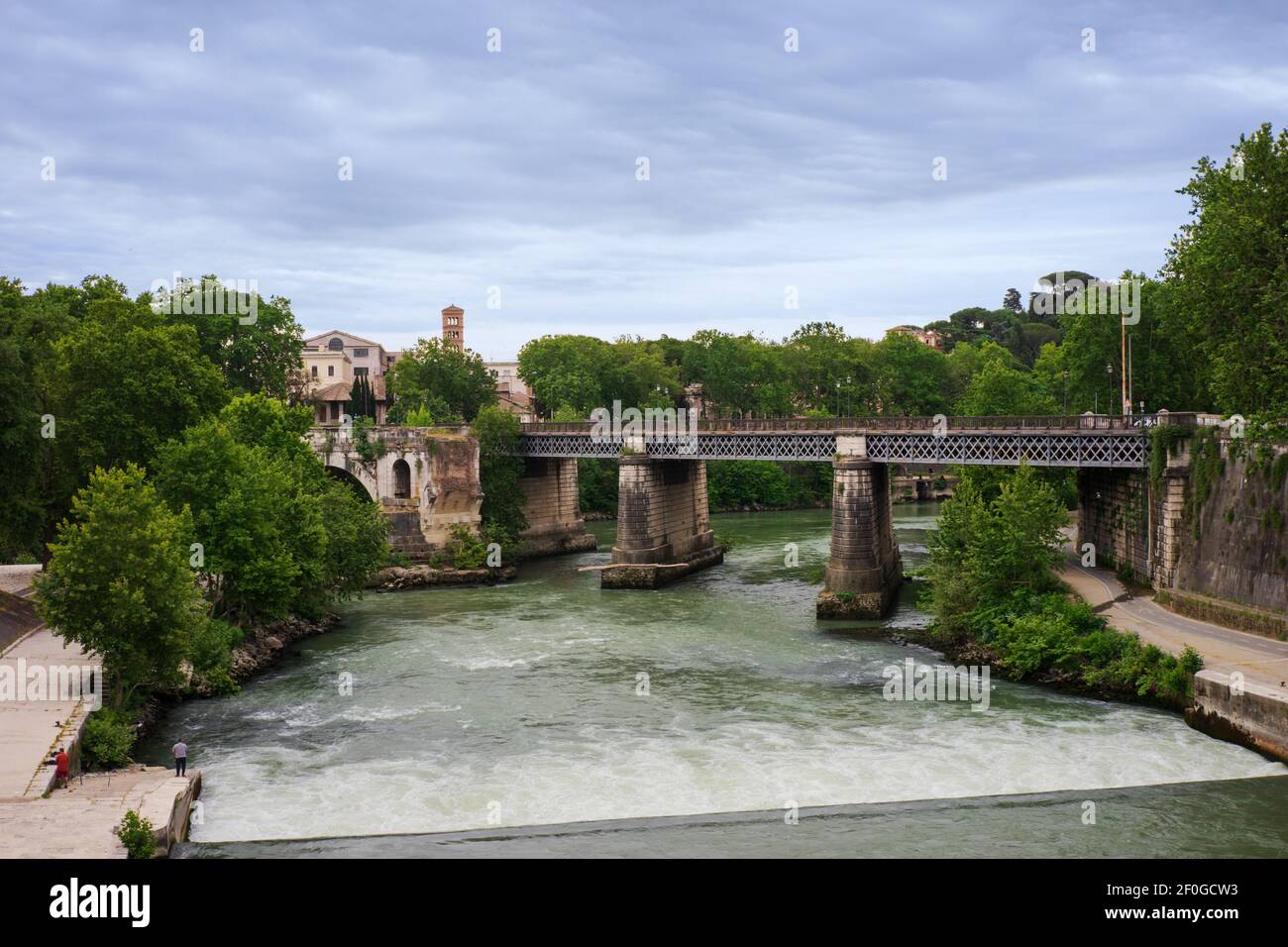 The water of the Tiber river flows under the Palatine bridge, lapping the old Ponte Rotto (Emilio) bridge, green trees surround the banks of the river Stock Photo