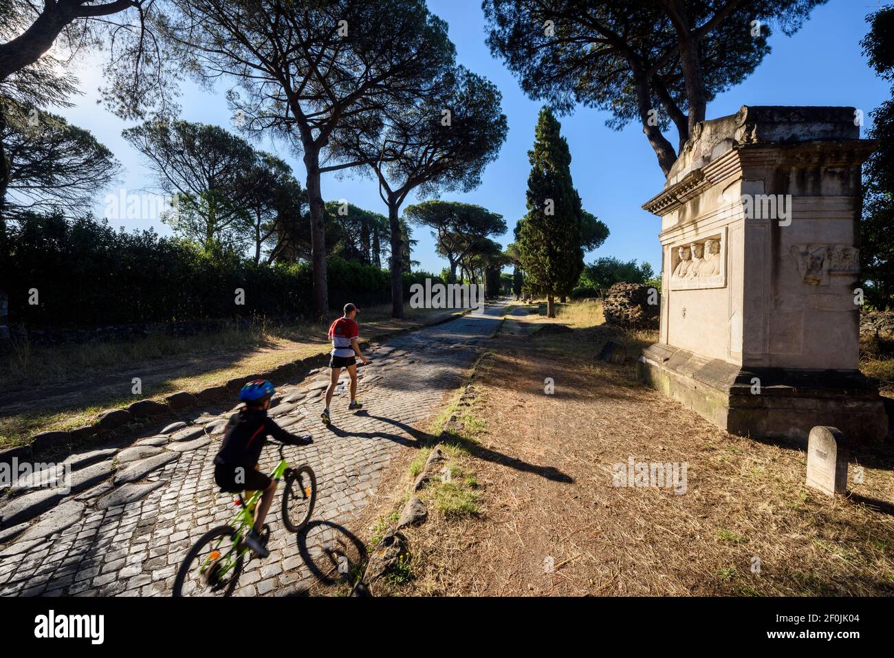 Rome. Italy. People running and cycling on the Via Appia Antica (Appian Way).  Right, ancient Roman Tomb of the Rabiri (Tomba dei Rabiri). Stock Photo