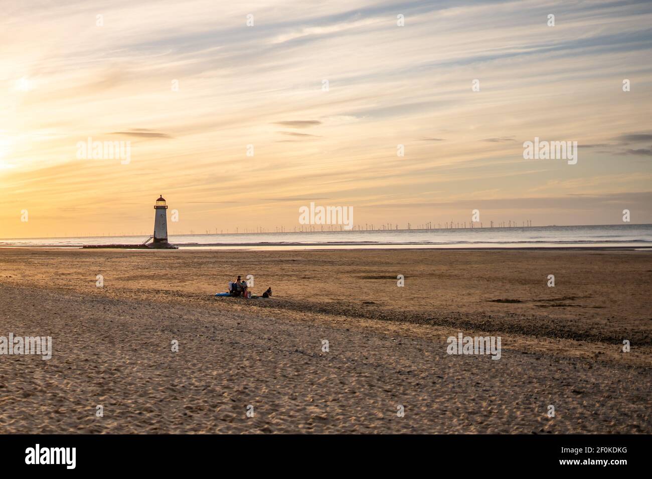 One family sat all alone together on deserted big clean beach vacation watching sunset with lighthouse and golden hour beautiful orange summer sky Stock Photo