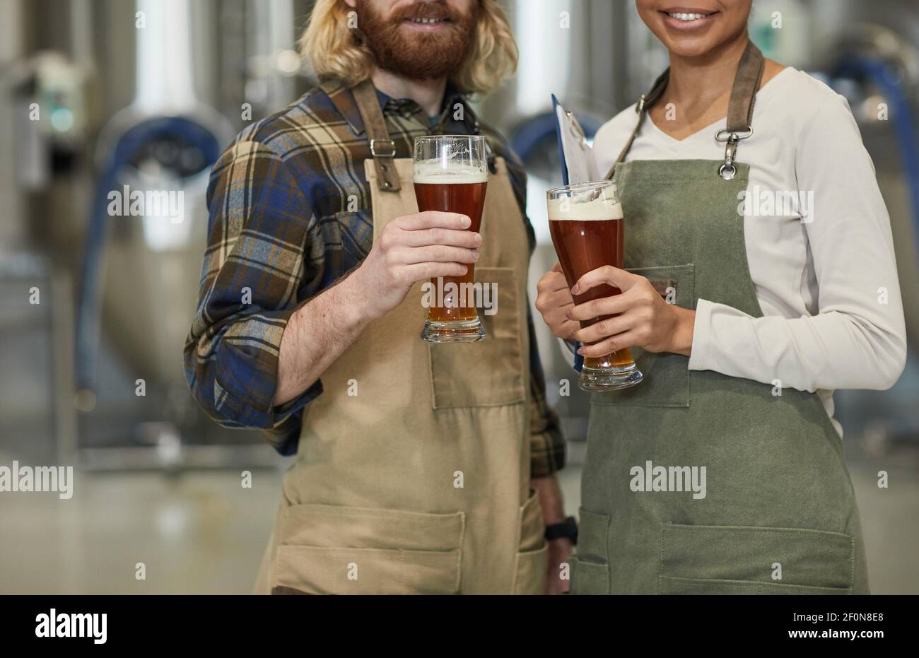 Cropped portrait of two smiling young workers holding beer glass and looking at camera while standing in workshop at brewing factory, copy space Stock Photo