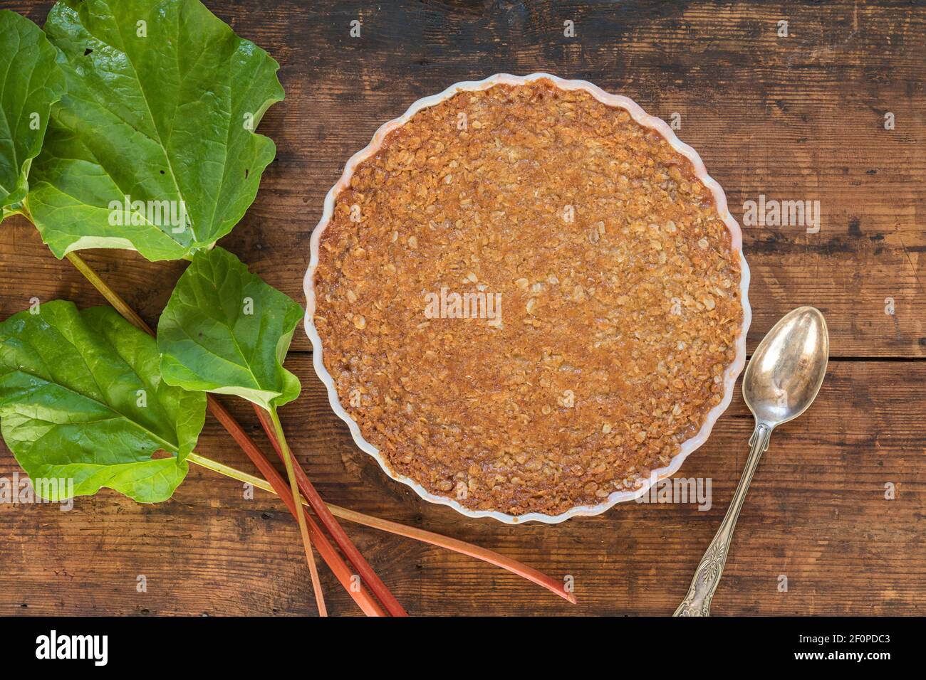 Traditional rhubarb pie on a rustic wooden table with a rhubarb stem. The pie is with a toffee topping made of rolled oats, cream, syrup, sugar, flour Stock Photo