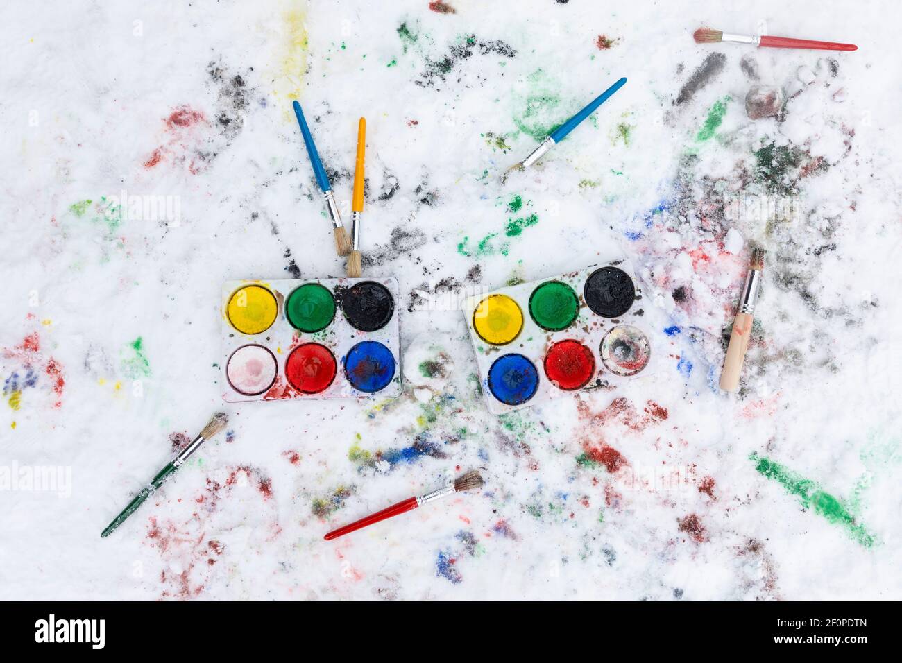 Watercolor painting on snow outdoors. Two water color paint boxes and scattered paint brushes seen from above. There is paint on the snow. Outdoors fu Stock Photo