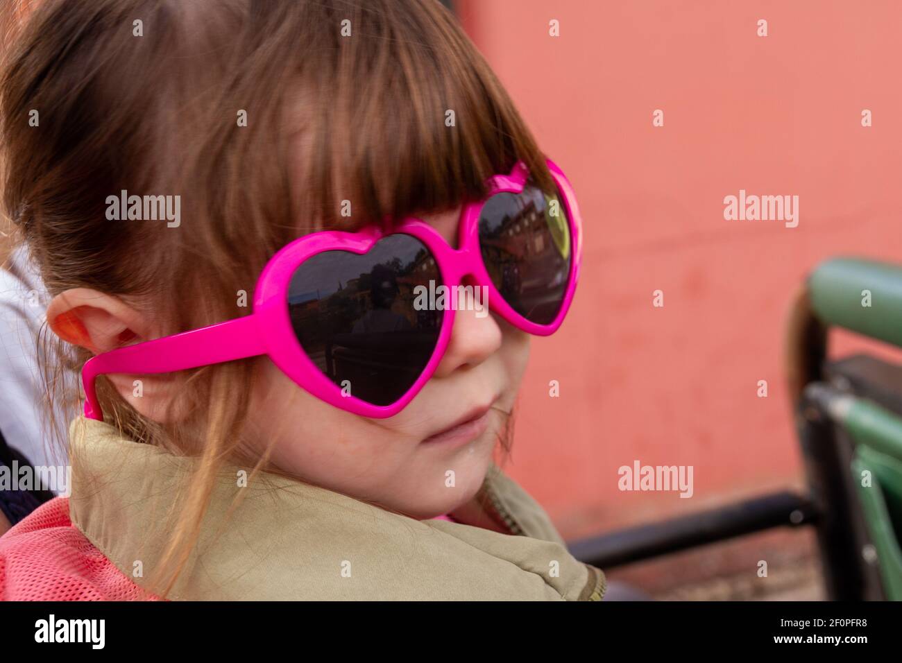 A cute, brown-haired baby girl wearing pink sunglasses and a green jacket Stock Photo