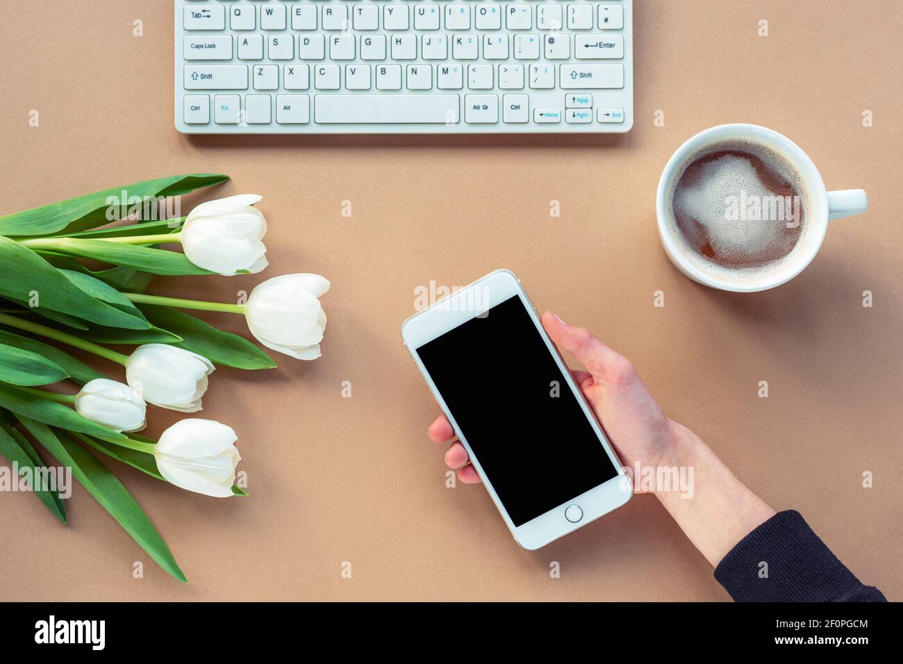 Workplace with coffee cup, hand holding a phone, computer keyboard and white tulips. Brown background. Women's day or Mother's day concept. Top view, Stock Photo