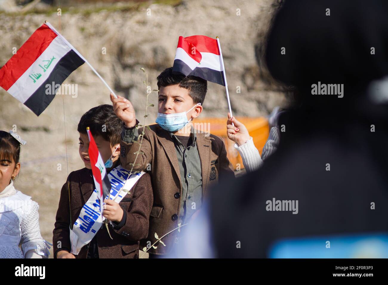 A kid waving the Iraqi flag during the papal visit. Pope Francis met the people and prayed for the souls of the war victims in Hosh al-Bieaa area near the ruins of the Al-Tahira Grand Church in the square which contains four churches all of which were damaged during the war against ISIS and this visit is considered the first papal visit in history to Iraq. Stock Photo